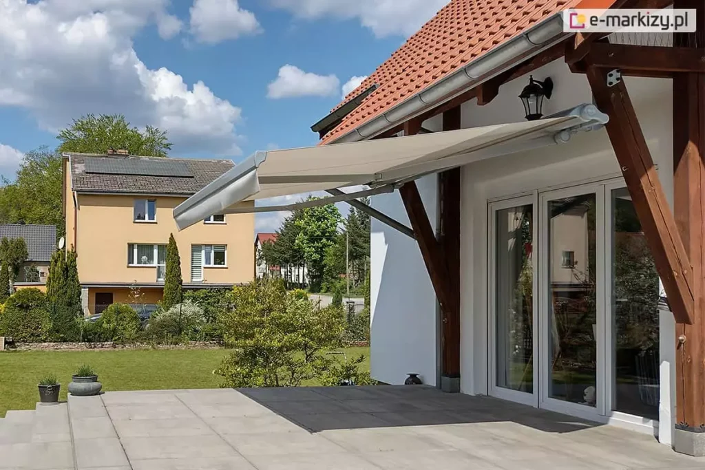 Zdjęcie prezentuje markizę tarasową w wersji bez kasetowej, zamontowaną w nietypowy sposób do drewnianych belek konstrukcyjnych (krokwi) zadaszenia tarasu. Markiza jest całkowicie rozłożona, a jej poszycie wykonane jest z gładkiej tkaniny w kolorze jasnobeżowym lub kremowym, zakończonej prostą falbaną. Aluminiowa konstrukcja markizy, w tym ramiona przegubowe i rura nawojowa, utrzymana jest w kolorze białym lub jasnoszarym. Osłona zacienia przestronny taras wyłożony dużymi płytami kamiennymi, znajdujący się przed białym domem z dwuskrzydłowymi, przeszklonymi drzwiami. Charakterystycznym elementem są masywne, ciemnobrązowe podpory drewniane, na których spoczywa mechanizm. W tle widoczny jest zadbany trawnik, sąsiedni budynek o żółtej elewacji z panelami fotowoltaicznymi na dachu oraz błękitne niebo z rozproszonymi chmurami.