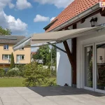 Zdjęcie prezentuje markizę tarasową w wersji bez kasetowej, zamontowaną w nietypowy sposób do drewnianych belek konstrukcyjnych (krokwi) zadaszenia tarasu. Markiza jest całkowicie rozłożona, a jej poszycie wykonane jest z gładkiej tkaniny w kolorze jasnobeżowym lub kremowym, zakończonej prostą falbaną. Aluminiowa konstrukcja markizy, w tym ramiona przegubowe i rura nawojowa, utrzymana jest w kolorze białym lub jasnoszarym. Osłona zacienia przestronny taras wyłożony dużymi płytami kamiennymi, znajdujący się przed białym domem z dwuskrzydłowymi, przeszklonymi drzwiami. Charakterystycznym elementem są masywne, ciemnobrązowe podpory drewniane, na których spoczywa mechanizm. W tle widoczny jest zadbany trawnik, sąsiedni budynek o żółtej elewacji z panelami fotowoltaicznymi na dachu oraz błękitne niebo z rozproszonymi chmurami.