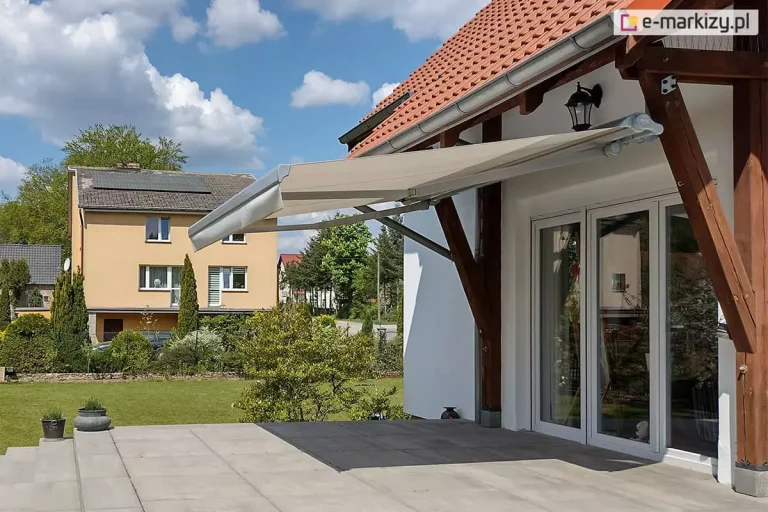 Zdjęcie prezentuje markizę tarasową w wersji bez kasetowej, zamontowaną w nietypowy sposób do drewnianych belek konstrukcyjnych (krokwi) zadaszenia tarasu. Markiza jest całkowicie rozłożona, a jej poszycie wykonane jest z gładkiej tkaniny w kolorze jasnobeżowym lub kremowym, zakończonej prostą falbaną. Aluminiowa konstrukcja markizy, w tym ramiona przegubowe i rura nawojowa, utrzymana jest w kolorze białym lub jasnoszarym. Osłona zacienia przestronny taras wyłożony dużymi płytami kamiennymi, znajdujący się przed białym domem z dwuskrzydłowymi, przeszklonymi drzwiami. Charakterystycznym elementem są masywne, ciemnobrązowe podpory drewniane, na których spoczywa mechanizm. W tle widoczny jest zadbany trawnik, sąsiedni budynek o żółtej elewacji z panelami fotowoltaicznymi na dachu oraz błękitne niebo z rozproszonymi chmurami.
