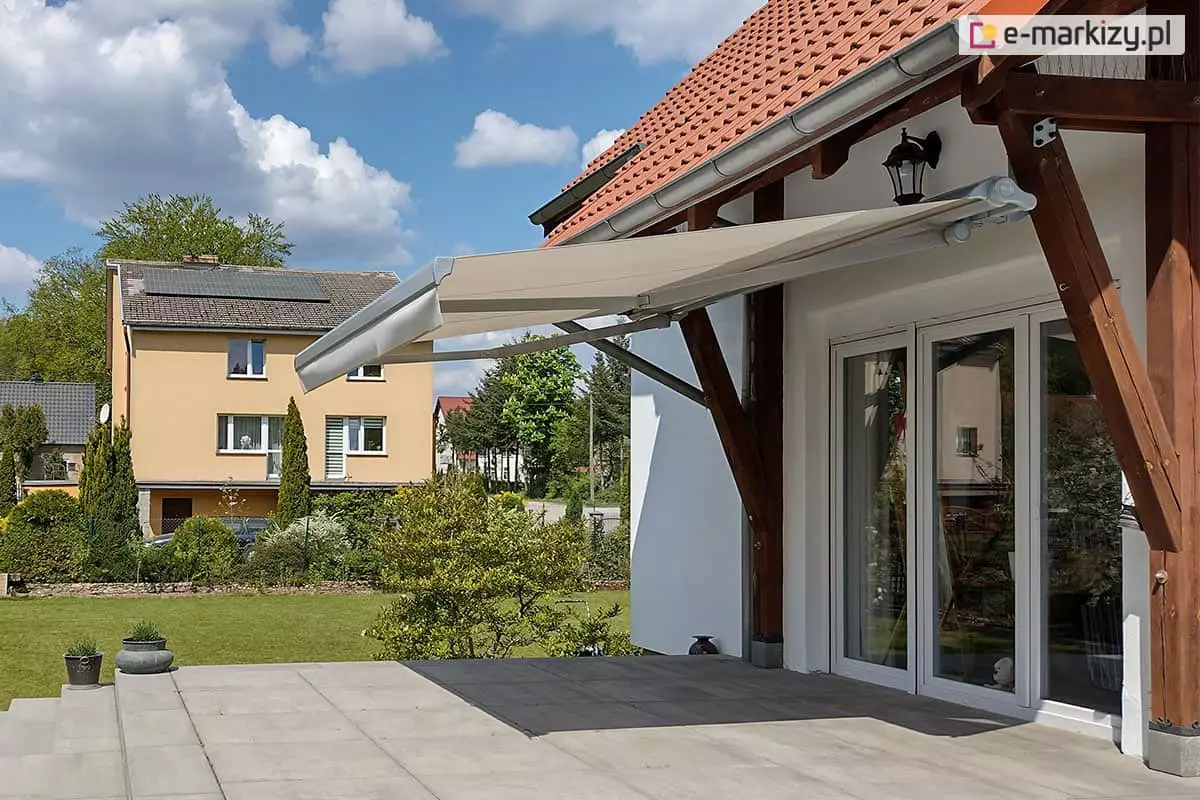 Zdjęcie prezentuje markizę tarasową w wersji bez kasetowej, zamontowaną w nietypowy sposób do drewnianych belek konstrukcyjnych (krokwi) zadaszenia tarasu. Markiza jest całkowicie rozłożona, a jej poszycie wykonane jest z gładkiej tkaniny w kolorze jasnobeżowym lub kremowym, zakończonej prostą falbaną. Aluminiowa konstrukcja markizy, w tym ramiona przegubowe i rura nawojowa, utrzymana jest w kolorze białym lub jasnoszarym. Osłona zacienia przestronny taras wyłożony dużymi płytami kamiennymi, znajdujący się przed białym domem z dwuskrzydłowymi, przeszklonymi drzwiami. Charakterystycznym elementem są masywne, ciemnobrązowe podpory drewniane, na których spoczywa mechanizm. W tle widoczny jest zadbany trawnik, sąsiedni budynek o żółtej elewacji z panelami fotowoltaicznymi na dachu oraz błękitne niebo z rozproszonymi chmurami.