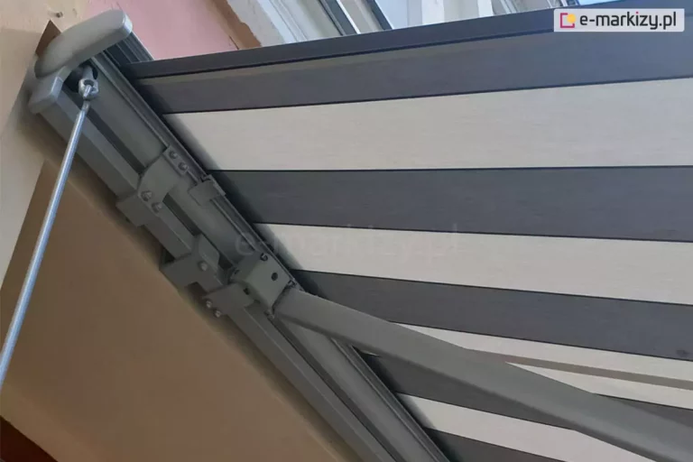Zbliżenie lewej strony markizy Palladio z grafitową konstrukcją oraz rozwiniętym poszyciem w pasy o trzech odcieniach szarości zamocowanej na elewacji nad wnęką okienną. Od boku widoczna zaślepka boczna z PCV oraz zaczep oczkowy przekładni korbowej do sterowania ręcznego, z zaczepioną o nią korbą. W głębi widoczne także, mocowania kasety, uchwyt ścienny oraz mocowanie ramienia markizy do belki kwadratowej.