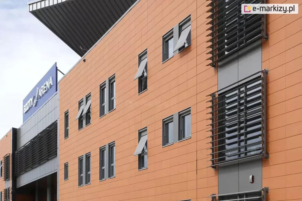 Zdjęcie przedstawia nowoczesną elewację budynku użyteczności publicznej (Netto Arena w Szczecinie) wykonaną z paneli fasadowych w kolorze pomarańczowym. Na ścianie zamontowano szereg pionowych, dwuskrzydłowych okien w szarych ramie, które wyposażono w systemy markizolet zewnętrznych. Tkanina osłon ma kolor jasnoszary, dopasowany do stolarki okiennej oraz elementów konstrukcyjnych budynku. Większość markizolet aluprof jest częściowo rozwinięta, a ich dolna sekcja została wychylona na zewnątrz pod kątem przy użyciu bocznych ramion, co tworzy charakterystyczne daszki nad dolną częścią przeszkleń. Na elewacji widoczne są również stałe łamacze światła w formie poziomych, ciemnych lameli zamontowanych na pionowych pasach fasady. W lewym górnym rogu zdjęcia widoczny jest fragment dachu oraz niebieski logotyp obiektu na szarym tle.