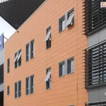 Zdjęcie przedstawia nowoczesną elewację budynku użyteczności publicznej (Netto Arena w Szczecinie) wykonaną z paneli fasadowych w kolorze pomarańczowym. Na ścianie zamontowano szereg pionowych, dwuskrzydłowych okien w szarych ramie, które wyposażono w systemy markizolet zewnętrznych. Tkanina osłon ma kolor jasnoszary, dopasowany do stolarki okiennej oraz elementów konstrukcyjnych budynku. Większość markizolet aluprof jest częściowo rozwinięta, a ich dolna sekcja została wychylona na zewnątrz pod kątem przy użyciu bocznych ramion, co tworzy charakterystyczne daszki nad dolną częścią przeszkleń. Na elewacji widoczne są również stałe łamacze światła w formie poziomych, ciemnych lameli zamontowanych na pionowych pasach fasady. W lewym górnym rogu zdjęcia widoczny jest fragment dachu oraz niebieski logotyp obiektu na szarym tle.