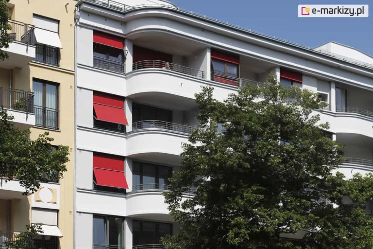 Zdjęcie prezentuje nowoczesny budynek mieszkalny o białej elewacji z zaokrąglonymi balkonami, na której zainstalowano systemy osłonowe w intensywnym czerwonym kolorze. Markizolety są zamontowane w niszach okiennych na kilku kondygnacjach, wprowadzając mocny akcent kolorystyczny do architektury obiektu. Osłony znajdują się w różnym stopniu rozwinięcia: niektóre są opuszczone do połowy wysokości okna z dolną częścią odchyloną pod kątem na zewnątrz, co tworzy charakterystyczny daszek przeciwsłoneczny. Inne pozostają niemal całkowicie zwinięte w górnych kasetach lub zasłaniają większą powierzchnię przeszklenia. Balkony wyposażone są w balustrady z poziomych metalowych prętów, a ich łukowaty kształt nadaje budynkowi dynamiczny wygląd. Po lewej stronie widoczny jest fragment sąsiedniego budynku w kolorze piaskowym z białymi osłonami okiennymi, a na pierwszym planie po prawej stronie rośnie gęste, zielone drzewo, które częściowo przesłania elewację. Całość uwieczniona została w pełnym świetle dziennym przy bezchmurnym, niebieskim niebie.