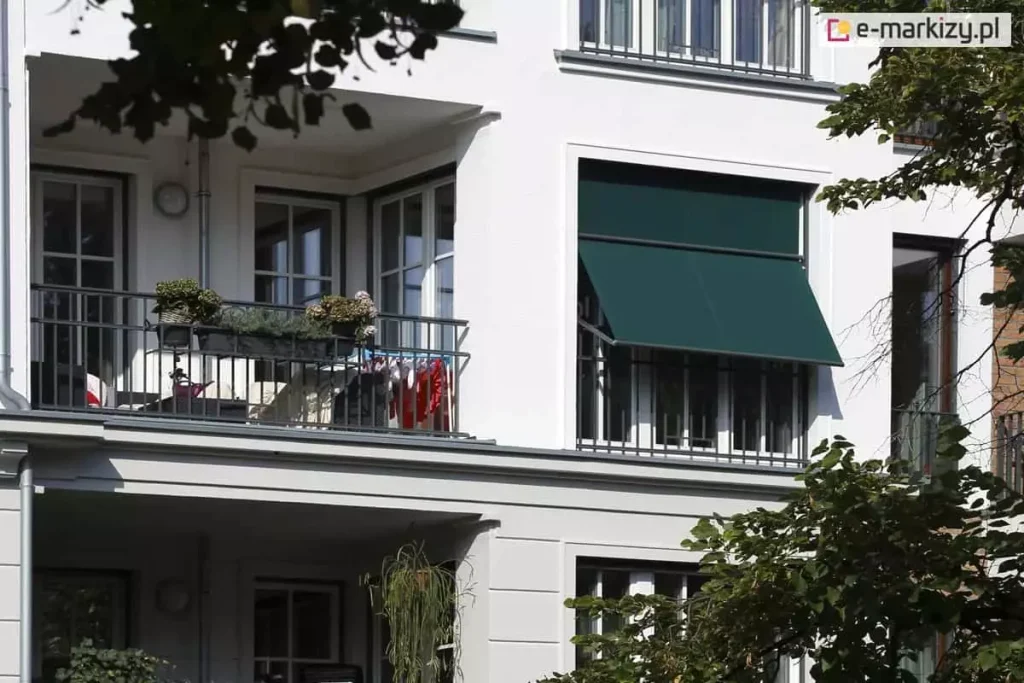Zdjęcie prezentuje jasną elewację budynku mieszkalnego z balkonami, na której zamontowano zewnętrzną markizoletę aluprof w kolorze ciemnej zieleni. Osłona zainstalowana jest w dużym otworze okiennym obok loggii i znajduje się w pozycji częściowo otwartej: górna część tkaniny przylega pionowo do okna, natomiast dolna sekcja jest odchylona na zewnątrz za pomocą bocznych ramion, tworząc daszek nad dolną częścią przeszklenia. Konstrukcja systemu, w tym kaseta i prowadnice, ma ciemny kolor dopasowany do barwy materiału. Obok, na sąsiednim balkonie z metalową balustradą, widoczne są donice z kwiatami oraz suszarka z praniem, co podkreśla użytkowy charakter osłony w codziennym życiu. W kadrze widoczne są również fragmenty zielonych liści drzew w narożnikach, a całość oświetlona jest naturalnym, ostrym światłem słonecznym, które rzuca wyraźne cienie na białą ścianę budynku.