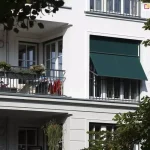 Zdjęcie prezentuje jasną elewację budynku mieszkalnego z balkonami, na której zamontowano zewnętrzną markizoletę aluprof w kolorze ciemnej zieleni. Osłona zainstalowana jest w dużym otworze okiennym obok loggii i znajduje się w pozycji częściowo otwartej: górna część tkaniny przylega pionowo do okna, natomiast dolna sekcja jest odchylona na zewnątrz za pomocą bocznych ramion, tworząc daszek nad dolną częścią przeszklenia. Konstrukcja systemu, w tym kaseta i prowadnice, ma ciemny kolor dopasowany do barwy materiału. Obok, na sąsiednim balkonie z metalową balustradą, widoczne są donice z kwiatami oraz suszarka z praniem, co podkreśla użytkowy charakter osłony w codziennym życiu. W kadrze widoczne są również fragmenty zielonych liści drzew w narożnikach, a całość oświetlona jest naturalnym, ostrym światłem słonecznym, które rzuca wyraźne cienie na białą ścianę budynku.