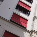 Zdjęcie prezentuje perspektywę z dołu na jasną elewację nowoczesnego budynku wielorodzinnego, na której zainstalowano systemy markizolet podtynkowych w intensywnym czerwonym kolorze. Osłony przeciwsłoneczne są wkomponowane w architekturę fasady, a ich kasety konstrukcyjne zostały ukryte pod warstwą tynku, co zapewnia minimalistyczny i estetyczny wygląd. Na fotografii widocznych jest kilka jednostek markizolet w różnym stopniu rozwinięcia: niektóre całkowicie przesłaniają otwory okienne w pionie, inne mają wysuniętą dolną część na ruchomych ramionach, tworząc charakterystyczny pochyły daszek. Taka funkcja pozwala na efektywne zacienienie wnętrza przy jednoczesnym zachowaniu widoku na zewnątrz i cyrkulacji powietrza. Czerwona tkanina akrylowa kontrastuje z kremową barwą ścian budynku oraz ciemnymi ramami okien i balustradami balkonowymi widocznymi w tle.