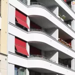 Zdjęcie przedstawia fragment nowoczesnej, białej elewacji budynku wielorodzinnego z charakterystycznymi, zaokrąglonymi balkonami. Na fasadzie zainstalowano systemy markizorolet zewnętrznych w intensywnym czerwonym kolorze, które pełnią funkcję osłon przeciwsłonecznych. Dwie markizorolety 120 aluprof po lewej stronie są częściowo rozwinięte, a ich dolna część została odchylona pod kątem za pomocą bocznych ramion, tworząc daszek osłaniający okna. Pozostałe osłony widoczne w głębi balkonów są w większości zwinięte lub opuszczone pionowo. Balkony posiadają balustrady wykonane z poziomych, metalowych rurek, a na niektórych z nich ustawione są skrzynki z biało-czerwonymi kwiatami. Po lewej stronie kadru widoczny jest fragment sąsiedniego budynku o beżowej elewacji z klasycznymi detalami architektonicznymi. Całość jest oświetlona mocnym, naturalnym światłem słonecznym, co tworzy wyraźne cienie na białych ścianach budynku.