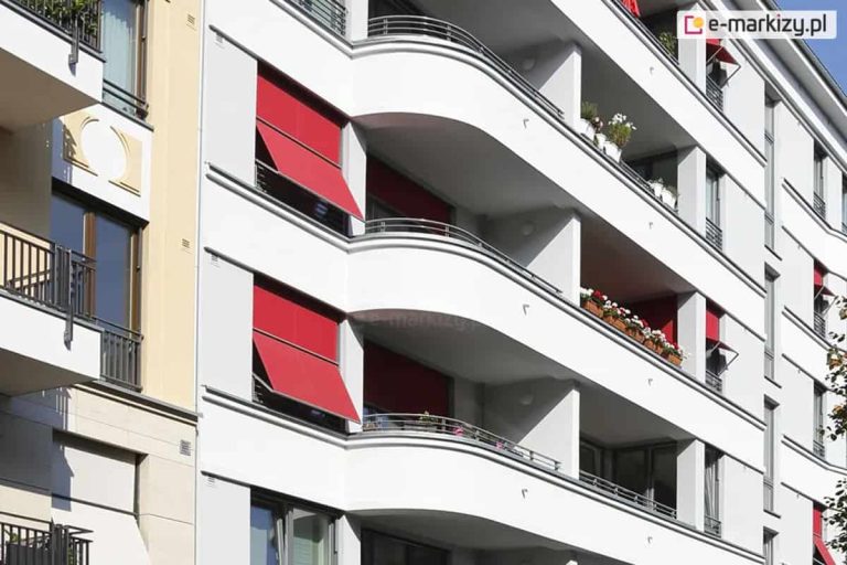 Zdjęcie przedstawia fragment nowoczesnej, białej elewacji budynku wielorodzinnego z charakterystycznymi, zaokrąglonymi balkonami. Na fasadzie zainstalowano systemy markizorolet zewnętrznych w intensywnym czerwonym kolorze, które pełnią funkcję osłon przeciwsłonecznych. Dwie markizorolety po lewej stronie są częściowo rozwinięte, a ich dolna część została odchylona pod kątem za pomocą bocznych ramion, tworząc daszek osłaniający okna. Pozostałe osłony widoczne w głębi balkonów są w większości zwinięte lub opuszczone pionowo. Balkony posiadają balustrady wykonane z poziomych, metalowych rurek, a na niektórych z nich ustawione są skrzynki z biało-czerwonymi kwiatami. Po lewej stronie kadru widoczny jest fragment sąsiedniego budynku o beżowej elewacji z klasycznymi detalami architektonicznymi. Całość jest oświetlona mocnym, naturalnym światłem słonecznym, co tworzy wyraźne cienie na białych ścianach budynku.