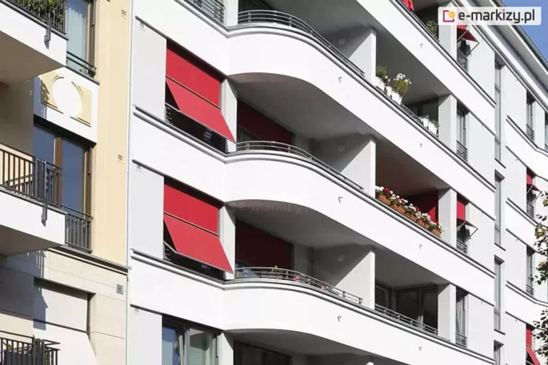 Zdjęcie przedstawia fragment nowoczesnej, białej elewacji budynku wielorodzinnego z charakterystycznymi, zaokrąglonymi balkonami. Na fasadzie zainstalowano systemy markizorolet zewnętrznych w intensywnym czerwonym kolorze, które pełnią funkcję osłon przeciwsłonecznych. Dwie markizorolety 120 aluprof po lewej stronie są częściowo rozwinięte, a ich dolna część została odchylona pod kątem za pomocą bocznych ramion, tworząc daszek osłaniający okna. Pozostałe osłony widoczne w głębi balkonów są w większości zwinięte lub opuszczone pionowo. Balkony posiadają balustrady wykonane z poziomych, metalowych rurek, a na niektórych z nich ustawione są skrzynki z biało-czerwonymi kwiatami. Po lewej stronie kadru widoczny jest fragment sąsiedniego budynku o beżowej elewacji z klasycznymi detalami architektonicznymi. Całość jest oświetlona mocnym, naturalnym światłem słonecznym, co tworzy wyraźne cienie na białych ścianach budynku.