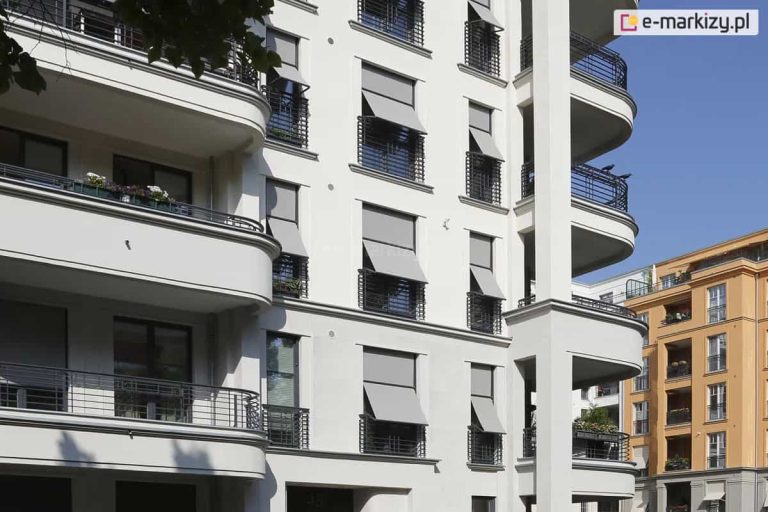 Zdjęcie ukazuje fragment jasnej elewacji wielopiętrowego budynku o klasycznej architekturze, na której zainstalowano systemy markizolet zewnętrznych w kolorze jasnoszarym. Osłony są zamontowane w niszach okiennych i charakteryzują się dwuetapowym sposobem rozwijania: górna część tkaniny przylega pionowo do płaszczyzny okna, natomiast dolna sekcja jest odchylona na stałych ramionach bocznych, tworząc funkcjonalny wysięg. Taka konstrukcja zapewnia skuteczne zacienienie wnętrza przy jednoczesnym zachowaniu swobodnego dopływu powietrza oraz widoku na zewnątrz przez dolną szczelinę. Na fasadzie widoczna jest powtarzalność montażu na różnych kondygnacjach, a systemy osłonowe są zintegrowane z estetyką budynku, współgrając z zaokrąglonymi balkonami i ciemnymi, metalowymi balustradami. W tle, po prawej stronie, widoczny jest fragment sąsiedniego budynku o pomarańczowej elewacji, co podkreśla miejski charakter zabudowy.