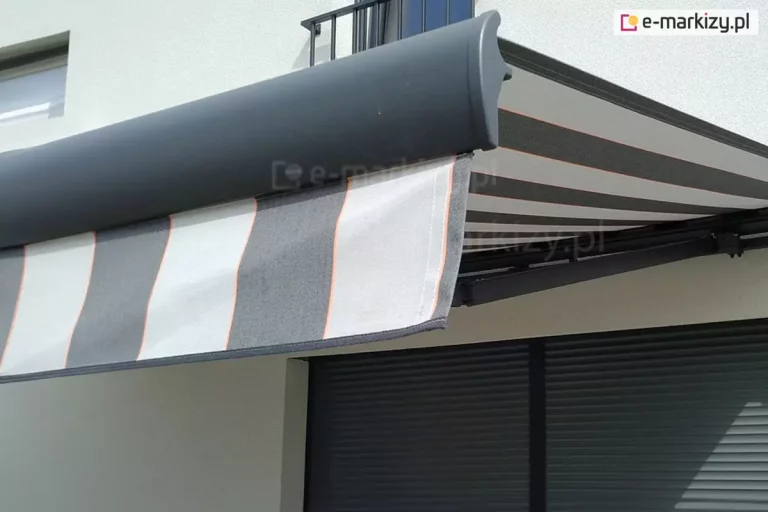 Markiza Palladio Selt w kolorze grafitowym z tkaniną w szare i jasnoszare pasy z cienkimi liniami w kolorze czerwonym zamocowana została na białej ścianie domu nad dużym oknem tarasowym przesłoniętym roletą zewnętrzną. Rozwinięte poszycie rzuca cień na ścianę i okno.