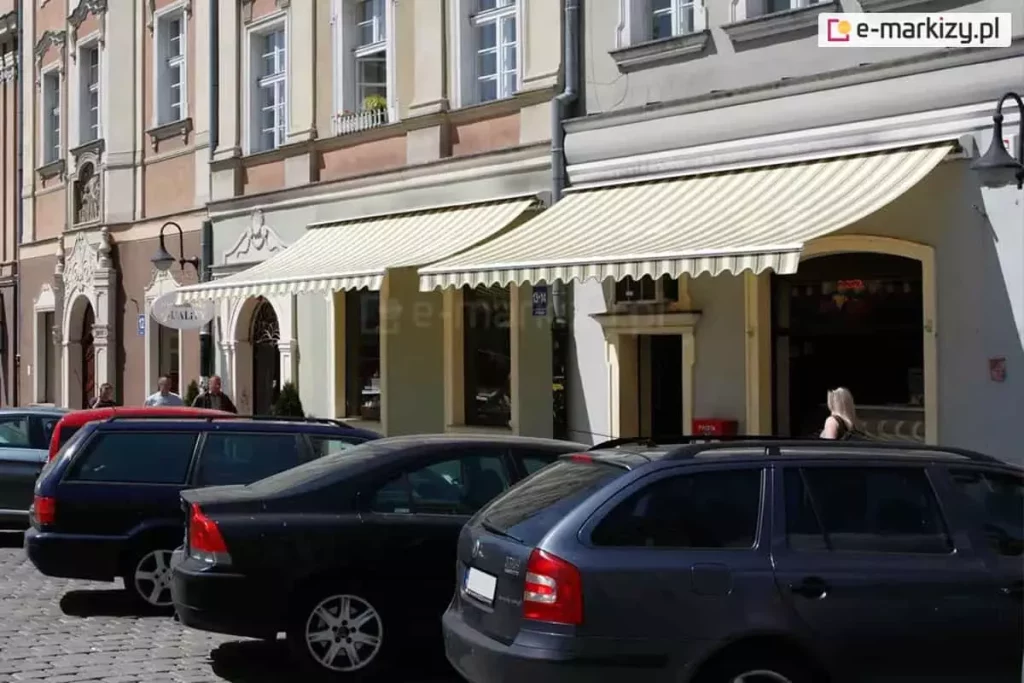 Duże witrynowe okna oraz wejście do sklepu mieszczącego się na parterze kamienicy osłonięte zostały dwoma umieszczonymi obok siebie markizami Jamaica Aluprof z uniwersalnym daszkiem nad tkaniną. Biała konstrukcja i tkanina w żółte i zielone pasy nie tylko skutecznie chronią przed słońcem, ale idealnie wpisują się w estetykę elewacji. Przed sklepem widoczne zaparkowane przy ulicy auta oraz kilku przechodniów.