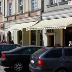 Duże witrynowe okna oraz wejście do sklepu mieszczącego się na parterze kamienicy osłonięte zostały dwoma umieszczonymi obok siebie markizami Jamaica Aluprof z uniwersalnym daszkiem nad tkaniną. Biała konstrukcja i tkanina w żółte i zielone pasy nie tylko skutecznie chronią przed słońcem, ale idealnie wpisują się w estetykę elewacji. Przed sklepem widoczne zaparkowane przy ulicy auta oraz kilku przechodniów.