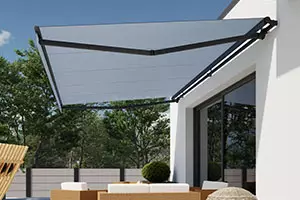 Rozwinięta markiza tarasowa z aluminiową konstrukcją w kolorze głębokiej czerni, zamontowana na bocznej, białej ścianie budynku. Rozpostarta tkanina markizy posiada jednolity, jasnoszary kolor i jest podtrzymywana przez składane ramiona przegubowe w kolorze czarnym, które zapewniają odpowiednie napięcie materiału oraz stabilność całej instalacji. Pod osłoną znajduje się wyodrębniona strefa wypoczynkowa z nowoczesnymi meblami z technorattanu, wyposażonymi w jasne poduszki siedziskowe, oraz niski stolik kawowy. W bezpośrednim sąsiedztwie strefy wypoczynkowej ustawiona jest dekoracyjna roślina w formie kuli oraz inne elementy małej architektury ogrodowej. Tło zdjęcia stanowi fragment przydomowego ogrodu z gęstym drzewostanem o zielonych liściach, widoczny za ogrodzeniem wykonanym z jasnych paneli. Całość sceny skąpana jest w pełnym świetle słonecznym, co podkreśla funkcjonalność markizy jako elementu zacieniającego taras i chroniącego przed bezpośrednim nasłonecznieniem w letnie dni.