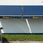 Dwie markizy sklepowe bez kasety Italia Selt zamocowane do białej ściany obok pomalowanej na niebiesko rynny. Dzięki nadrukowi na logo na falbanie, firma, której siedzibę zdobią markizy zyskała dobrą reklamę przy parkingu widocznym obok skrawka zielonego trawnika. Po lewej widoczna część parkującego auta marki Daewoo.