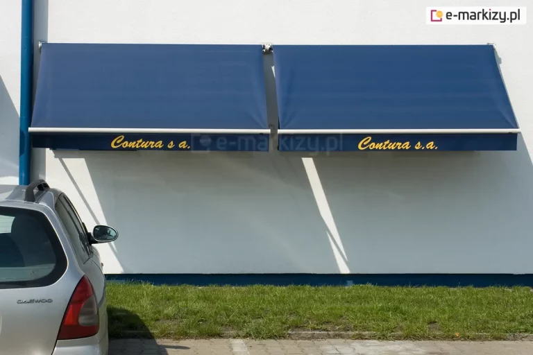 Dwie markizy sklepowe bez kasety Italia Selt zamocowane do białej ściany obok pomalowanej na niebiesko rynny. Dzięki nadrukowi na logo na falbanie, firma, której siedzibę zdobią markizy zyskała dobrą reklamę przy parkingu widocznym obok skrawka zielonego trawnika. Po lewej widoczna część parkującego auta marki Daewoo.