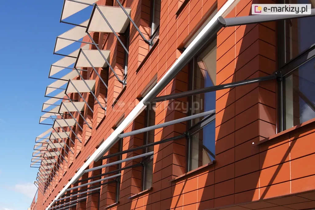 Elewacja nowoczesnego budynku biurowego lub użyteczności publicznej wykonana z ceramicznych płyt elewacyjnych w kolorze ceglastym o gładkiej strukturze i wyraźnych poziomych podziałach. Na fasadzie zamontowano system markiz sklepowych Italia bez kasety z długimi ramionami mocowanymi do ramy okien. Markizy mają formę prostokątnych paneli z jasnej, półtransparentnej tkaniny w odcieniu beżowym, rozpiętych na metalowych konstrukcjach ramowych. Każda markiza jest zamocowana pod kątem do lica ściany za pomocą ciemnoszarych, stalowych wysięgników i prowadnic, co nadaje elewacji trójwymiarowy, geometryczny rytm. Widok ujęty jest z perspektywy żabiej, patrząc w górę wzdłuż ściany budynku, co podkreśla powtarzalność elementów osłonowych na kolejnych kondygnacjach. W tle widoczne jest bezchmurne, intensywnie błękitne niebo, a ostre światło słoneczne rzuca wyraźne, ukośne cienie ram markiz na pomarańczową elewację, akcentując teksturę materiałów i architektoniczne detale systemu montażowego.