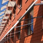 Elewacja nowoczesnego budynku biurowego lub użyteczności publicznej wykonana z ceramicznych płyt elewacyjnych w kolorze ceglastym o gładkiej strukturze i wyraźnych poziomych podziałach. Na fasadzie zamontowano system markiz sklepowych Italia bez kasety z długimi ramionami mocowanymi do ramy okien. Markizy mają formę prostokątnych paneli z jasnej, półtransparentnej tkaniny w odcieniu beżowym, rozpiętych na metalowych konstrukcjach ramowych. Każda markiza jest zamocowana pod kątem do lica ściany za pomocą ciemnoszarych, stalowych wysięgników i prowadnic, co nadaje elewacji trójwymiarowy, geometryczny rytm. Widok ujęty jest z perspektywy żabiej, patrząc w górę wzdłuż ściany budynku, co podkreśla powtarzalność elementów osłonowych na kolejnych kondygnacjach. W tle widoczne jest bezchmurne, intensywnie błękitne niebo, a ostre światło słoneczne rzuca wyraźne, ukośne cienie ram markiz na pomarańczową elewację, akcentując teksturę materiałów i architektoniczne detale systemu montażowego.
