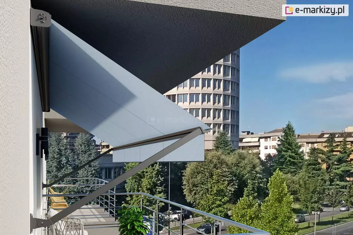 Zdjęcie prezentuje zbliżenie od boku na konstrukcję markizy balkonowej zamontowanej na jasnej elewacji budynku wielorodzinnego. Markiza o jasnoszarym poszyciu jest częściowo wysunięta, chroniąc przestrzeń balkonu przed słońcem. Widoczne są solidne, ciemnoszare ramiona boczne mocowane bezpośrednio do ściany za pomocą metalowych uchwytów, które stabilizują materiał. Górna część mechanizmu znajduje się pod krawędzią wyższej kondygnacji, co zapewnia dodatkową ochronę dla samej kasety urządzenia. Na balkonie widoczna jest nowoczesna balustrada z połyskujących, poziomych profili metalowych, za którą znajduje się fragment plecionego fotela oraz donica z gęstą, zieloną roślinnością. W tle rozpościera się widok na miejską panoramę, w tym na charakterystyczny wysoki budynek o cylindrycznym kształcie z licznymi oknami, otoczony przez korony drzew i niższe bloki mieszkalne. Perspektywa zdjęcia została ujęta z boku, co pozwala dokładnie przyjrzeć się kątowi nachylenia materiału markizy oraz elementom montażowym przytwierdzonym do białego tynku budynku.