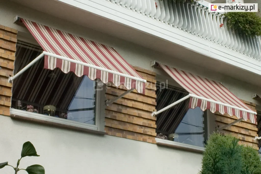 Dwie markizy bez kasety Italia Aluprof zamocowane nad oknami budynku z jasną fasadą oraz zdobieniami z cegieł pomiędzy oknami. Białe ramiona markiz zamocowane zostały do ściany w pobliży wnęki okiennej, a poszycie w bordowo, biało, brązowe pasy i falowana falbana zapewniają zacienienie pomieszczeniom za oknami. Powyżej markiz widoczna balustrada betonowa balkonu, a pod oknem z prawej zielone krzaki.