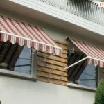 Dwie markizy bez kasety Italia Aluprof zamocowane nad oknami budynku z jasną fasadą oraz zdobieniami z cegieł pomiędzy oknami. Białe ramiona markiz zamocowane zostały do ściany w pobliży wnęki okiennej, a poszycie w bordowo, biało, brązowe pasy i falowana falbana zapewniają zacienienie pomieszczeniom za oknami. Powyżej markiz widoczna balustrada betonowa balkonu, a pod oknem z prawej zielone krzaki.
