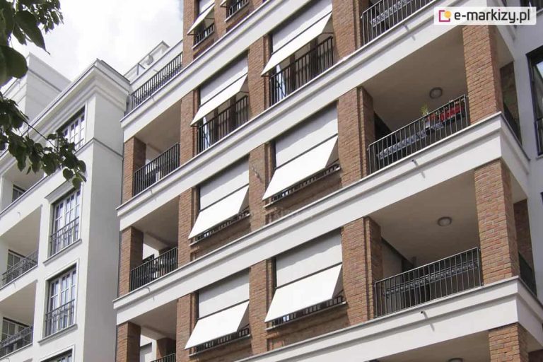 Zdjęcie przedstawia nowoczesny budynek wielorodzinny o elewacji wykonanej z czerwonej cegły z wyraźnymi białymi elementami konstrukcyjnymi i szerokimi gzymsami. Na balkonach zamontowano pionowe markizy zewnętrzne typu screen w jasnym, niemal białym kolorze. Osłony te są częściowo rozwinięte, przy czym ich dolna część jest odchylona na zewnątrz za pomocą bocznych ramion, co tworzy charakterystyczny daszek zapewniający dopływ powietrza przy jednoczesnym zacienieniu wnętrza. Balustrady balkonowe są ażurowe, wykonane z ciemnego metalu w formie pionowych szczebli. Markizorolety 103 są zamontowane w niszach okienno-balkonowych, tworząc uporządkowany i estetyczny rytm na fasadzie budynku. W tle widoczne jest jasne, lekko zachmurzone niebo oraz fragment sąsiedniego, białego budynku o klasycznej architekturze z dużymi oknami i czarnymi barierkami.