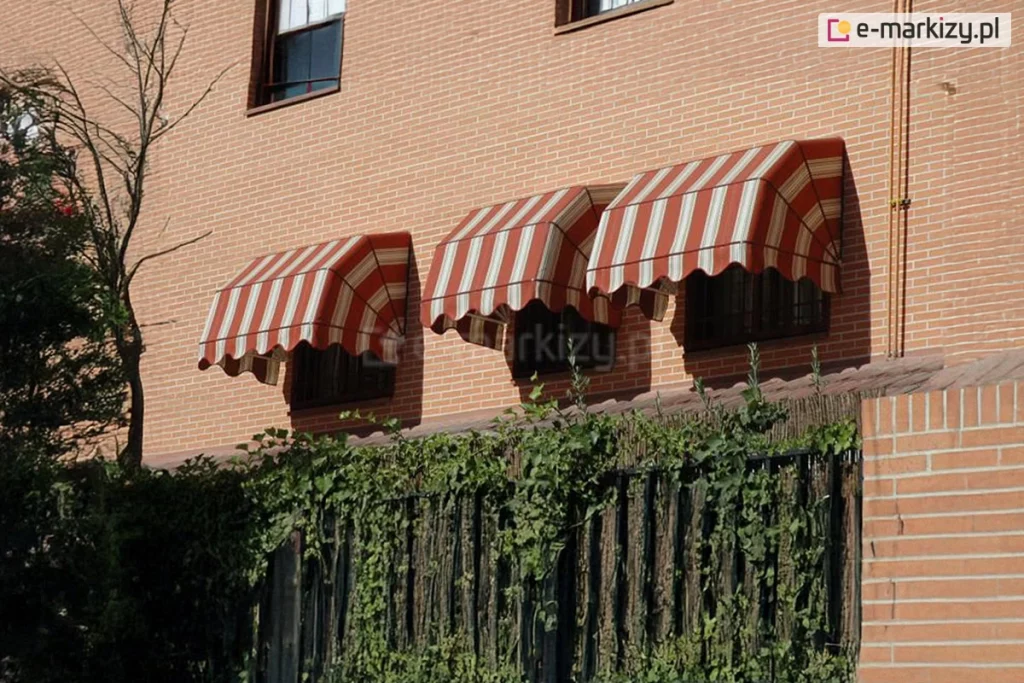 Zdjęcie prezentuje efekt końcowy montażu trzech markiz koszowych na elewacji budynku wykonanej z jasnobrązowej cegły klinkierowej. Markizy o klasycznym, prostym kształcie zostały zainstalowane bezpośrednio nad otworami okiennymi. Poszycie markiz posiada wzór w pionowe pasy w kolorach kremowym i czerwonym, a dolna krawędź wykończona jest dekoracyjną, pofalowaną falbaną z lamówką. Konstrukcje są w pełni rozłożone, osłaniając okna przed słońcem. W tle widoczny jest fragment ogrodu z zieloną roślinnością pnącą się po ogrodzeniu oraz fragment sąsiedniego budynku.