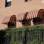 Zdjęcie prezentuje efekt końcowy montażu trzech markiz koszowych na elewacji budynku wykonanej z jasnobrązowej cegły klinkierowej. Markizy o klasycznym, prostym kształcie zostały zainstalowane bezpośrednio nad otworami okiennymi. Poszycie markiz posiada wzór w pionowe pasy w kolorach kremowym i czerwonym, a dolna krawędź wykończona jest dekoracyjną, pofalowaną falbaną z lamówką. Konstrukcje są w pełni rozłożone, osłaniając okna przed słońcem. W tle widoczny jest fragment ogrodu z zieloną roślinnością pnącą się po ogrodzeniu oraz fragment sąsiedniego budynku.