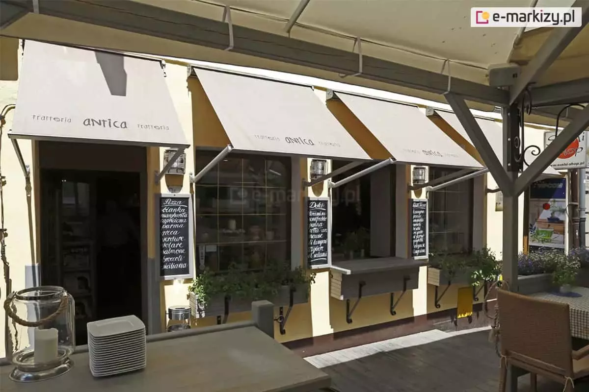 Cztery markizy balkonowo sklepowe Italia kasetowa z ramionami mocowanymi do ściany oraz nadrukiem na poszyciu markizy tuż nad belką dolną osłaniają wejście oraz trzy okna kawiarni z ogródkiem dla klientów umieszczonym pod zadaszoną konstrukcją.
