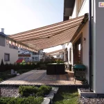 Szeroka osłona tarasowa w poziome, brązowo-beżowe pasy, rozłożona nad przydomowym miejscem do wypoczynku. Poszycie wykończone jest dekoracyjną, pofalowaną falbaną z ciemniejszą lamówką i zamontowane na elewacji piętrowego, jasnego budynku. Pod zadaszeniem znajduje się duży, ciemny stół ogrodowy oraz grill ustawiony przy ścianie domu. Taras został wyłożony kostką brukową w dwóch odcieniach, a jego krawędź wyznaczają niskie, betonowe palisady. Wokół strefy wypoczynkowej rosną niskie krzewy iglaste posadzone w szarym żwirze, a w tle widać rząd wysokich tui oddzielających posesję od sąsiednich, nowoczesnych budynków. W tle widoczne są sąsiednie ogrody zabudowy szeregowej osłonięte identycznymi markizami. Po lewej stronie, w głębi ogrodu, za metalowym płotem, widoczny jest mały, kolorowy domek dla dzieci. Całość kompozycji oświetla mocne, popołudniowe słońce, rzucając wyraźny cień osłony na nawierzchnię tarasu.