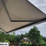 Zdjęcie przedstawia nowoczesną markizę tarasową Moderno w pełnej kasecie, widoczną od strony dolnej mechanizmu oraz poszycia. Konstrukcja opiera się na dwóch solidnych, przegubowych ramionach bocznych wykonanych z metalu o matowym wykończeniu w kolorze ciemnoszarym lub antracytowym. Ramiona te podtrzymują rozciągniętą tkaninę poszycia w kolorze jasnokremowym lub piaskowym, która jest napięta i tworzy jednolitą płaszczyznę osłaniającą przestrzeń przed słońcem. Na krawędzi zewnętrznej widoczna jest estetyczna, prostokątna listwa przednia o ostrych, nowoczesnych liniach, która po złożeniu markizy szczelnie domyka kasetę. W tle znajduje się ogród z gęstą roślinnością, drzewami liściastymi oraz fragmentem domu jednorodzinnego o żółtej elewacji i ciemnym dachu, a nad całością rozciąga się lekko zachmurzone niebo.