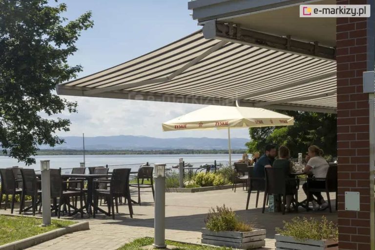Zdjęcie prezentuje rozłożystą markizę Corsica zamontowaną na tarasie restauracji lub kawiarni z widokiem na jezioro i góry. Tkanina markizy jest wykończona w klasyczne, szerokie pasy w kolorze białym i szarym, co nadaje miejscu elegancki, a zarazem wypoczynkowy charakter. Konstrukcja markizy Corsica, widoczna w górnej części kadru, opiera się na solidnych ramionach, które pozwalają na zacienienie znacznej powierzchni tarasu. Pod zadaszeniem znajdują się meble ogrodowe wykonane z ciemnego technorattanu – stoły oraz krzesła, przy których siedzą goście lokalu. Taras jest wyłożony jasną kostką brukową, a na jego obrzeżach ustawiono niskie, drewniane skrzynie z roślinnością oraz nowoczesne lampy ogrodowe w formie słupków. W tle, poza obrębem markizy, widoczny jest duży, biały parasol gastronomiczny z logo marki piwa. Najdalszy plan zdjęcia ukazuje taflę jeziora z cumującymi łodziami oraz zamglone pasmo górskie pod pogodnym niebem. Po prawej stronie kadr zamyka fragment elewacji budynku wykonanej z ciemnoczerwonej cegły klinkierowej. Całość kompozycji podkreśla funkcjonalność markizy Corsica w przestrzeniach komercyjnych, zapewniającej komfort termiczny i ochronę przed słońcem w miejscach rekreacyjnych.