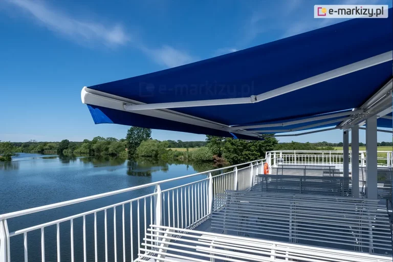 Zdjęcie przedstawia duży, otwarty pokład pasażerski statku lub barki, który został osłonięty systemem nowoczesnych markiz tarasowych. Dominującym elementem jest intensywnie niebieskie, gładkie poszycie markiz, które rozciąga się nad rzędami białych, metalowych ławek o konstrukcji szczebelkowej. Stelaż markiz oraz barierki ochronne statku są polakierowane na kolor biały, co kontrastuje z barwą materiału i błękitem nieba. Widoczne są dwa rzędy ramion przegubowych, które stabilizują zadaszenie. W tle rozpościera się malowniczy krajobraz rzeczny – spokojna tafla wody odbija błękitne niebo oraz gęstą zieloną roślinność porastającą brzegi rzeki. Na horyzoncie widoczna jest linia drzew pod czystym, słonecznym niebem. Perspektywa zdjęcia ujęta z poziomu pokładu podkreśla dużą powierzchnię zacienienia uzyskaną dzięki zastosowaniu profesjonalnego systemu osłonowego w warunkach nasłonecznienia.