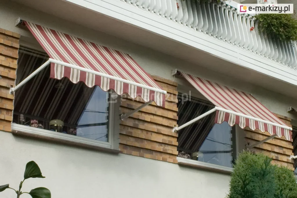 Dwie markizy bez kasety Italia Selt zamocowane nad oknami budynku z jasną fasadą oraz zdobieniami z cegieł pomiędzy oknami. Białe ramiona markiz zamocowane zostały do ściany w pobliży wnęki okiennej, a poszycie w bordowo, biało, brązowe pasy i falowana falbana zapewniają zacienienie pomieszczeniom za oknami. Powyżej markiz widoczna balustrada betonowa balkonu, a pod oknem z prawej zielone krzaki.