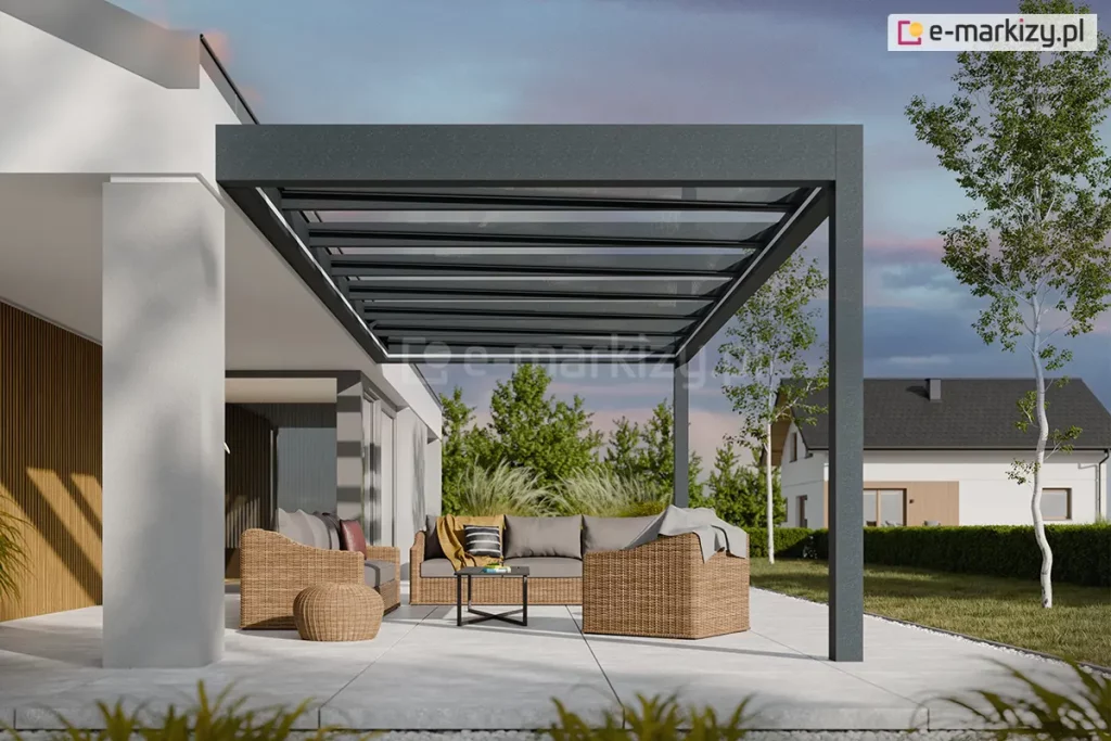 Wizualizacja przedstawia nowoczesną realizację zadaszenia tarasu Spekta Glass, zintegrowaną z bryłą minimalistycznego, jasnego budynku jednorodzinnego. Konstrukcja nośna została wykonana z aluminiowych profili w kolorze antracytowym, które tworzą ramę dachu zamontowaną jednostronnie do elewacji i wspartą na zewnętrznych, pionowych słupach. Dach konstrukcji jest w całości przeszklony i podzielony na regularne sekcje, co zapewnia pełną przezierność i ochronę przed warunkami atmosferycznymi. Pod zadaszeniem zaaranżowano strefę wypoczynkową z meblami z technorattanu w odcieniu naturalnego brązu, obejmującą sofy, fotele oraz niską pufę. Podłoże tarasu stanowią wielkoformatowe płyty tarasowe w kolorze jasnoszarym, które płynnie łączą się z zadbanym trawnikiem. W tle widoczna jest nowoczesna architektura ogrodowa, niska roślinność oraz sąsiednie budynki, co podkreśla spójność konstrukcji z otoczeniem.
