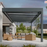 Wizualizacja przedstawia nowoczesną realizację zadaszenia tarasu Spekta Glass, zintegrowaną z bryłą minimalistycznego, jasnego budynku jednorodzinnego. Konstrukcja nośna została wykonana z aluminiowych profili w kolorze antracytowym, które tworzą ramę dachu zamontowaną jednostronnie do elewacji i wspartą na zewnętrznych, pionowych słupach. Dach konstrukcji jest w całości przeszklony i podzielony na regularne sekcje, co zapewnia pełną przezierność i ochronę przed warunkami atmosferycznymi. Pod zadaszeniem zaaranżowano strefę wypoczynkową z meblami z technorattanu w odcieniu naturalnego brązu, obejmującą sofy, fotele oraz niską pufę. Podłoże tarasu stanowią wielkoformatowe płyty tarasowe w kolorze jasnoszarym, które płynnie łączą się z zadbanym trawnikiem. W tle widoczna jest nowoczesna architektura ogrodowa, niska roślinność oraz sąsiednie budynki, co podkreśla spójność konstrukcji z otoczeniem.