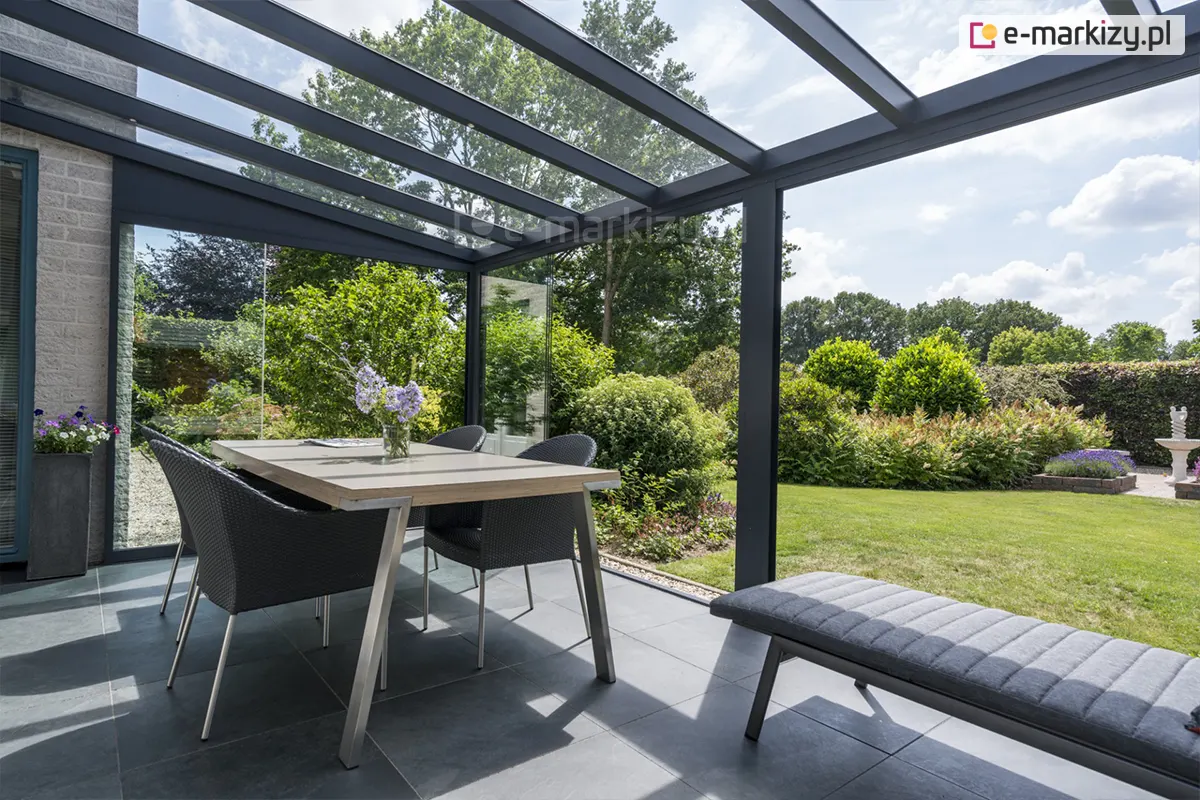 Zdjęcie prezentuje perspektywę z wnętrza nowoczesnego zadaszenia tarasowego model Terano, ukazującą strefę jadalnianą oraz wypoczynkową zintegrowaną z ogrodem. Konstrukcja zadaszenia opiera się na profilach aluminiowych w kolorze antracytowym, z dachem wypełnionym przezroczystymi panelami szklanymi, które zapewniają swobodny dostęp światła dziennego. Po lewej stronie widoczne jest stałe przeszklenie boczne, zamontowane przy elewacji budynku z jasnej cegły, przy której stoi wysoka, ciemna donica z kwiatami. Centralnym punktem aranżacji jest prostokątny stół z jasnym blatem na metalowym stelażu, przy którym ustawiono cztery wygodne fotele z technorattanu w kolorze czarnym. Na pierwszym planie po prawej stronie znajduje się fragment nowoczesnej ławki lub pufy z szarym, pikowanym siedziskiem. Podłoga tarasu wyłożona jest dużymi płytami w kolorze ciemnoszarym. Otwarta konstrukcja otwiera widok na zadbany ogród z zielonym trawnikiem, formowanymi krzewami, paprociami oraz elementem małej architektury w postaci fontanny widocznej w oddali.