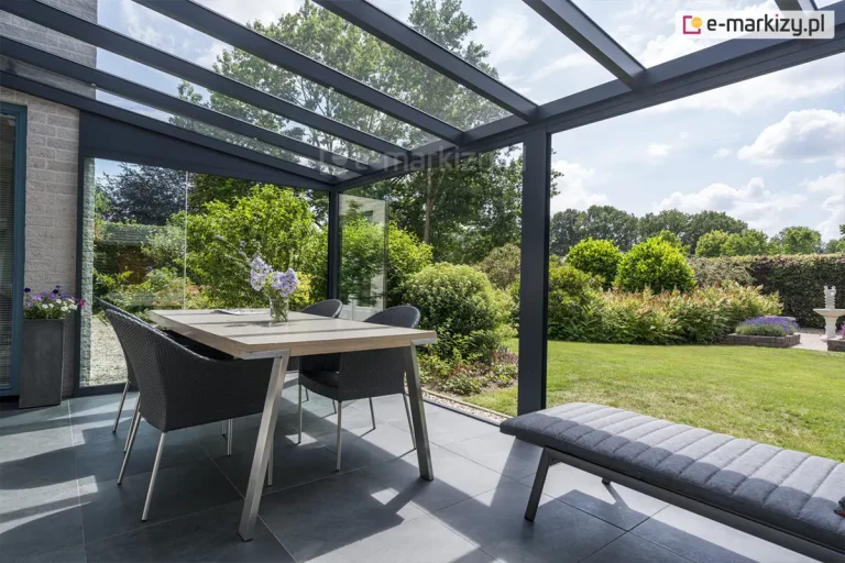 Nowoczesna aranżacja strefy jadalnianej na tarasie zadaszonym systemem aluminiowo-szklanym w kolorze antracytowym. Konstrukcja dachu składa się z przezroczystych paneli szklanych opartych na równoległych krokwiach, wspartych na pionowym słupie o kwadratowym przekroju. Pod zadaszeniem ustawiono stół z jasnym blatem na metalowych nogach, przy którym znajdują się cztery fotele z ciemnej plecionki technorattanowej. W dolnej części kadru widoczny jest fragment tapicerowanej ławy lub leżanki w kolorze szarym. Posadzka tarasu została wykonana z wielkoformatowych płyt gresowych w kolorze ciemnoszarym. Zabudowa posiada boczne przeszklenia stałe lub przesuwne, które oddzielają wnętrze od ogrodu. W tle roztacza się widok na zadbany ogród z bujną roślinnością, krzewami ozdobnymi oraz trawnikiem pod słonecznym niebem. Przy ścianie budynku z jasnej cegły ustawiono wysoką, ciemną donicę z kwitnącymi kwiatami.