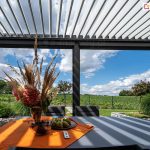 Stół jadalny z bukietem kwiatów zacieniony przez uchylone lamele pergoli SB400 z widocznym w tle sielski, wiejskim otoczeniem i niebieskim niebem