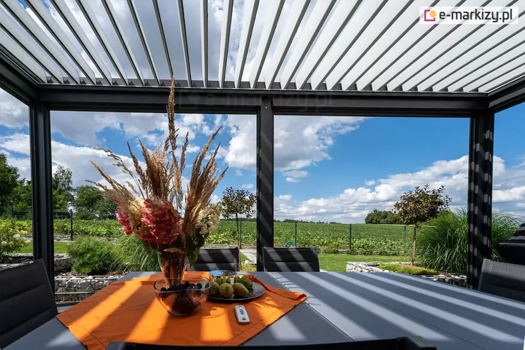 Wnętrze nowoczesnej pergoli tarasowej Sb400 o konstrukcji aluminiowej w kolorze antracytowym, wyposażonej w dach z ruchomych lameli (bioklimatyczny). Lamele są częściowo otwarte, przepuszczając pasy światła słonecznego na stół. Pod zadaszeniem znajduje się szary stół ogrodowy z pomarańczowym bieżnikiem, na którym stoi wazon z suszonymi trawami i kwiatami, miska z ciemnymi owocami oraz talerz z gruszkami. Obok owoców leży biały pilot do sterowania automatyką pergoli. W tle widoczny jest ogród z zieloną roślinnością, niskimi drzewkami oraz rozległe pole pod zachmurzonym, niebieskim niebem. Konstrukcja posiada pionowe profile wsporcze, a podłoże wokół tarasu wykończone jest gabionami wypełnionymi jasnym kamieniem.
