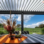 Wnętrze nowoczesnej pergoli tarasowej Sb400 o konstrukcji aluminiowej w kolorze antracytowym, wyposażonej w dach z ruchomych lameli (bioklimatyczny). Lamele są częściowo otwarte, przepuszczając pasy światła słonecznego na stół. Pod zadaszeniem znajduje się szary stół ogrodowy z pomarańczowym bieżnikiem, na którym stoi wazon z suszonymi trawami i kwiatami, miska z ciemnymi owocami oraz talerz z gruszkami. Obok owoców leży biały pilot do sterowania automatyką pergoli. W tle widoczny jest ogród z zieloną roślinnością, niskimi drzewkami oraz rozległe pole pod zachmurzonym, niebieskim niebem. Konstrukcja posiada pionowe profile wsporcze, a podłoże wokół tarasu wykończone jest gabionami wypełnionymi jasnym kamieniem.