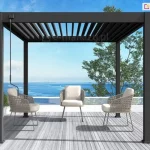 Wolnostojąca pergola lamelowa Marlo w kolorze antracytowym, zainstalowana na nowoczesnym tarasie z widokiem na morze. Konstrukcja o kwadratowym przekroju słupów posiada dach wykonany z ruchomych lameli aluminiowych, które są ustawione w pozycji częściowo otwartej. Taras wyłożony jest deskami kompozytowymi w kolorze jasnoszarym i zabezpieczony szklaną balustradą z białą ramą, co zapewnia nieograniczony widok na błękitną wodę oraz horyzont. Pod zadaszeniem znajduje się minimalistyczna strefa wypoczynkowa składająca się z trzech foteli o ażurowym, beżowym splocie z białymi poduszkami siedziskowymi. Przy lewym frontowym słupku konstrukcyjnym zamontowana jest czarna korba służąca do ręcznej regulacji kąta nachylenia dachu. W tle, po prawej stronie, widoczne są zielone korony drzew liściastych na tle czystego, błękitnego nieba.