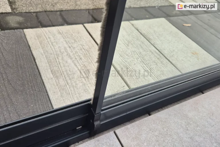 Zbliżenie dwóch mijających się paneli szklanych bezramowych MIROS zamocowanych w aluminiowej antracytowej prowadnicy. Na krawędzi prawego panelu zamocowano mocowaną w aluminiowej listwie nasuwanej na szkło uszczelkę szczotkową, która chroni przed wiatrem i owadami.