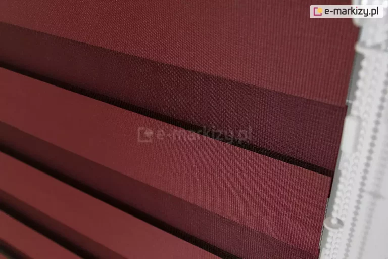 Zbliżenie tkaniny dzień i noc do rolet. Wyraźnie widoczne dwie warstwy tkaniny z ułożonymi naprzemiennie pasami w kolorze bordo oraz przejrzystym. Odpowiednie ustawienie pasów tkaniny zapewnia pełne zakrycie szyby. Po prawej widoczny łańcuszek do sterowania roletką w kolorze białym.