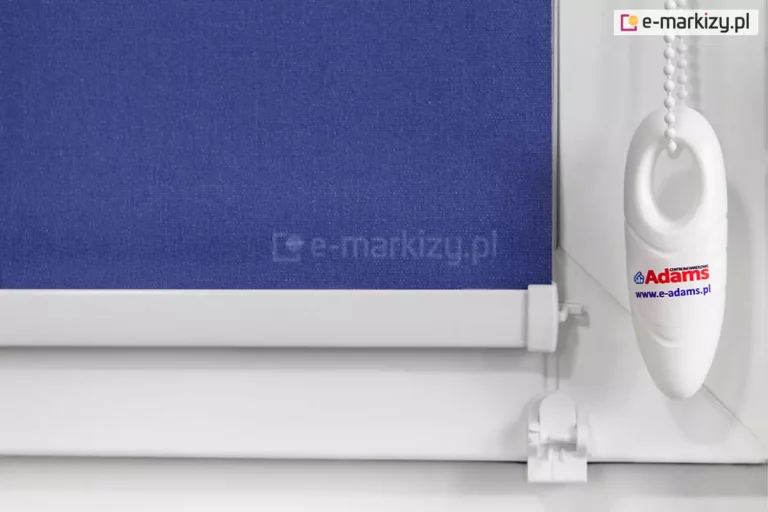 Ujęcie szczegółów rolety mini: obciążnika łańcuszka z naklejką producenta, mocowania prowadnicy żyłkowej do ramy okna, niebieskiej tkaniny oraz aluminiowego obciążnika tkaniny. Całość zamocowana do okna o białej ramie.