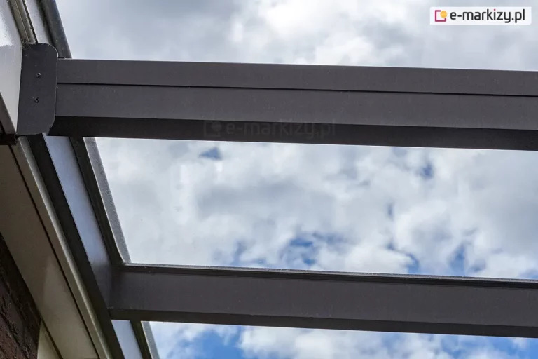 Zbliżenie na mocowanie konstrukcji oraz krokwi dachowych zadaszenia Terano w listwie instalowanej do ściany. Pomiędzy krokwiami widoczne przez szkło zachmurzone niebo.