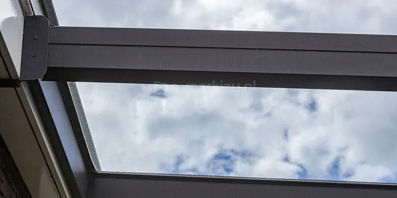 Zbliżenie listwy ściennej zadaszenia Terano, do której mocowane są antracytowe krokwie, pomiędzy którymi, przez przejrzyste tafle szkła widoczne jest zachmurzone niebo.