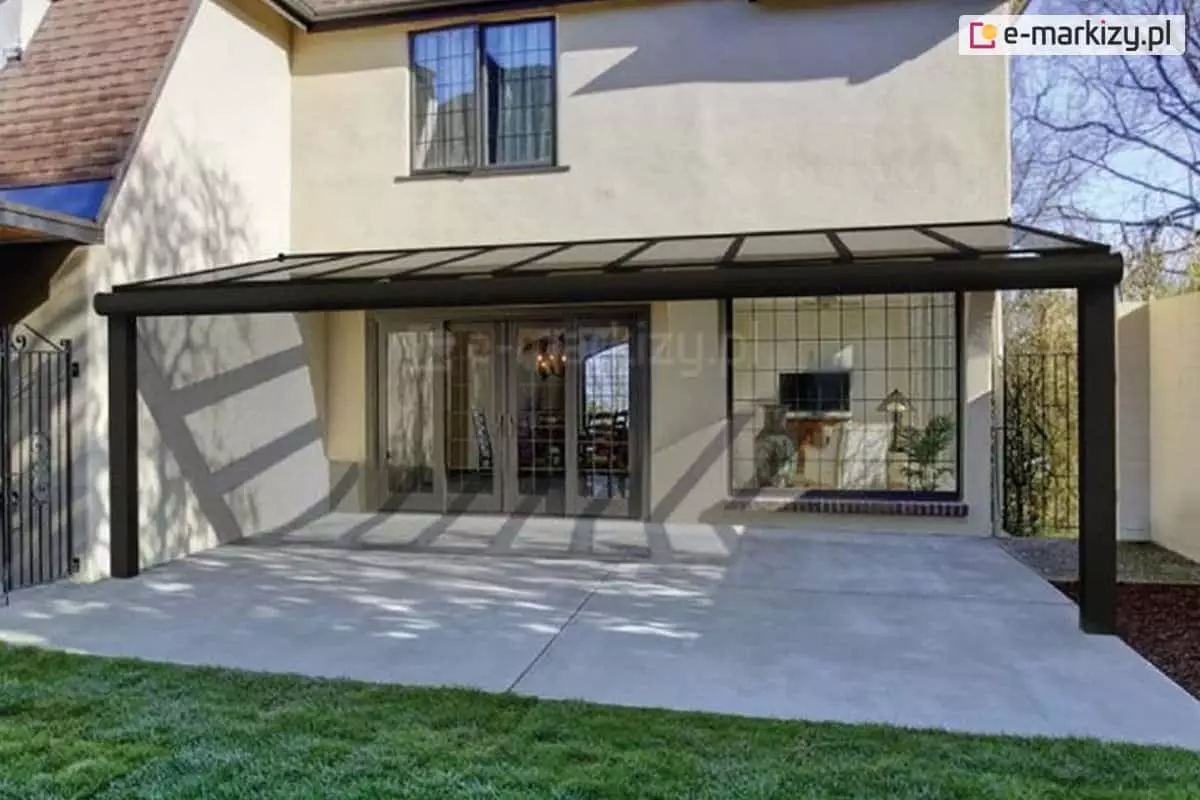 Nowoczesne zadaszenie tarasowe Modes Glas o ciemnej, antracytowej konstrukcji, zamontowane do jasnej elewacji piętrowego domu jednorodzinnego. Jednospadowy dach zadaszenia wypełniony jest dużymi, przezroczystymi taflami szkła, które są podtrzymywane przez równolegle rozmieszczone, smukłe krokwie. Całość opiera się na dwóch solidnych, pionowych słupach nośnych usytuowanych na krawędziach tarasu, przy czym przednia belka poprzeczna posiada estetyczne, zaokrąglone wykończenie rynny. Pod zadaszeniem, przy jasnej, betonowej wylewce tarasu, widoczne są szerokie, przeszklone drzwi tarasowe z ozdobnymi szprosami oraz duże okno, przez które dostrzec można stylowe oświetlenie wnętrza salonu. Na elewacji budynku widoczna jest gra świateł i cieni rzucanych przez konstrukcję, a po lewej stronie znajduje się ozdobna, metalowa furtka prowadząca do ogrodu z zielonym trawnikiem.