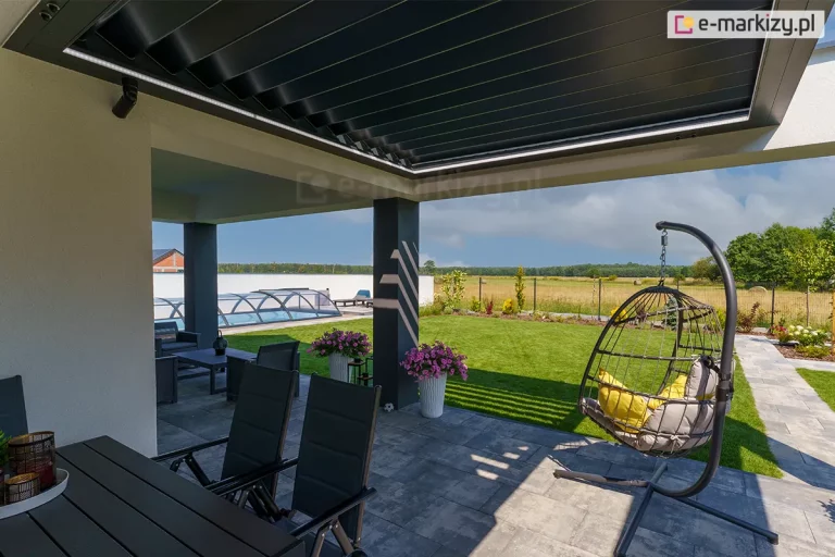 Zdjęcie prezentuje perspektywę z wnętrza tarasu skierowaną na ogród, ukazującą bioklimatyczny dach lamelowy SB400R w pozycji lekko uchylonej. Aluminiowe lamele w kolorze antracytowym tworzą jednolitą płaszczyznę zadaszenia, chroniącą przed słońcem i deszczem. Wzdłuż wewnętrznej krawędzi ramy konstrukcyjnej zamontowano liniowe oświetlenie LED, które emituje jasne, białe światło, doświetlając przestrzeń pod pergolą. Konstrukcja wsparta jest na masywnym, ciemnoszarym słupie nośnym, a posadzka tarasu wykonana jest z dużych, jasnoszarych płyt betonowych. W strefie wypoczynkowej widoczny jest wiszący fotel ogrodowy typu kokon z szarymi poduszkami oraz czarne krzesła przy stole jadalnianym. W tle rozpościera się widok na zadbany ogród z zielonym trawnikiem, basenem przykrytym niską, transparentną kopułą oraz otwartą przestrzeń pól pod błękitnym niebem z rozproszonymi chmurami.